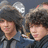 Two pixelated figures with dark hair and light skin wear dark jackets over blue shirts against a gray background.