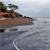 Pixelated static image of a brown boat on gray water with green foliage on the left, under a light blue sky.