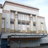 Pixelated static image of a multi-story building with rectangular windows and a flat roof, set against a light sky background.