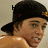 Pixelated image of a person wearing a black cap with visible text and a yellow-and-red emblem, shirtless against a light background.