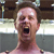 Pixelated close-up of a person with light skin and short brown hair, mouth open in a shout or yell, against a blurred background with horizontal light and dark bands.