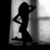 Black-and-white silhouette of a woman in a long dress standing near a window with one hand on her head and the other on her hip.