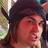 Pixelated close-up of a person with dark hair wearing a dark cap, face turned slightly right against blurred background.