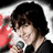 Pixelated image of a person with dark hair holding a microphone, wearing a white shirt and black jacket against a light background.