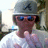 A person wearing a gray cap with a black symbol, blue sunglasses, and a white shirt, making a hand gesture with fingers extended.