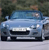 Static image of a light blue sedan with a dark grille and headlights, positioned on a light gray surface against a blurred green background.