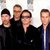Four pixelated figures in dark clothing stand closely together against a light background, with one figure wearing a blue jacket.