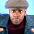 Static pixelated image of a person wearing a flat cap and dark jacket over maroon shirt against light blue background.
