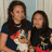 Two people with long dark hair stand side-by-side holding a small dog wearing a red and white outfit against a light-colored wall background.