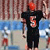 Pixelated static image of a football player in black uniform with red helmet and number 6 on chest, standing against blurred background with blue water and brown structures.