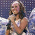 A woman with long brown hair holds a microphone, wearing a white top and brown pants with dark blue jeans underneath, standing against a starry background with white light effects.