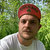 Pixelated image of a person wearing a red head covering and light-colored shirt against green foliage background.