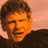 Pixelated static image of a person's face with brown hair and orange-toned skin against a light peach background, wearing dark clothing.