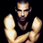 Pixelated static image of a man with short hair and beard wearing a black sleeveless shirt, arms crossed against dark background.