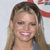 A pixelated close-up of a person with light brown hair tied back, wearing a blue garment, and smiling with visible teeth against a light background.