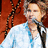 Pixelated image of a person in light blue clothing holding a microphone against a red background.