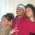 Three people pose together in a red and beige outfit with a headband, against a light background.