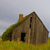 Pixelated static image of a small brown house with a green roof, black door, and chimney, set against a light gray sky and yellow grass.