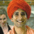 Two individuals wearing red turbans and light-colored shirts stand side-by-side with a third person partially visible behind them, all smiling against a blurred background.