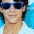 A person wearing blue sunglasses and a white shirt against a bright blue background, with another figure in grayscale nearby.