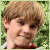 Pixelated static image of a person with light brown hair and green foliage background, cropped to show upper face and shoulders.