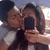 Two people pose for a selfie with one holding a black smartphone, the background shows blurred buildings and light.