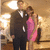 A couple in formal attire stands in a warmly lit indoor setting with a blurred background, the woman in a pink dress and the man in a dark suit.