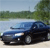 A blue sedan with visible headlights and wheels is positioned on a light-colored surface, set against a blurred background of greenery and sky.