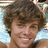 Pixelated close-up of a person with curly brown hair, light skin, and a smiling expression against a blurred background.