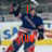 A hockey player in a dark blue jersey and red pants skates on ice with a stick, surrounded by blurred arena advertisements.