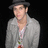 Pixelated image of a person wearing a dark fedora, light gray jacket over white shirt with visible buttons and colorful patterned tie against black background.