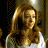 A pixelated image of a person with long light brown hair wearing a white top, set against a dark patterned background with a bright light source on the right.