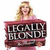 A woman with long blonde hair in a pink dress holds a maroon sign reading LEGALLY BLONDE in silver letters, smiling at the camera.