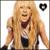 Pixelated image of a person with long blonde hair wearing a light jacket over dark clothing, holding a microphone against a bright background.