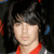 A person with dark shoulder-length hair and bangs wears a red-collared shirt under a gray jacket with white stripes, shown in four with subtle facial expression changes