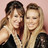 Two women with long hair pose closely together against a light pink background, one in dark clothing and the other in black sleeves.