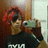 A person with red hair holds a silver camera, wearing a black shirt with white text, against a brown and white background with pink text overlay.