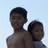 Two shirtless individuals stand close together against a light blue background, one slightly behind the other, both facing forward with neutral expressions.