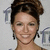 A woman with brown hair in an updo smiles at the camera against a light background with repeated 'T' logos.
