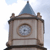 Pixelated clock tower with a blue spire, circular clock face, and brown stone-like structure against a light blue sky background.