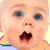 Pixelated close-up of a baby's face with wide blue eyes, open mouth showing dark interior, and light skin tone against yellow background.