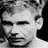 Black-and-white pixelated close-up of a person's face with visible hair and ear, rendered in low-resolution grayscale with dithering effects on the background.