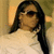 A person with long dark hair and sunglasses wears a white high-necked top against a blurred background.