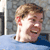 Pixelated close-up of a person with light brown hair, visible ear, and mouth open in a smile against a blurred background.