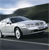 A silver sedan with dark tinted windows is positioned on a road, viewed from a low angle with a blurred background of sky and hills.