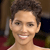 Pixelated close-up of a person with short dark hair, light skin, and a broad smile showing teeth, wearing a sleeveless top against a blurred background with brown and red tones.