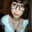 Pixelated static image of a person with dark hair wearing a light blue top, pink lips, and a dark necklace against a brown background.