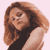 Pixelated static image of a person with long brown hair wearing a black top and light-colored outer garment against a pale pink background.