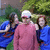 Group of people in outdoor setting with greenery background; one person wears pink sweater and white cap, others wear blue tops.