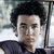 A person with dark curly hair wearing a dark sleeveless top, seated inside a vehicle with a blurred outdoor background.